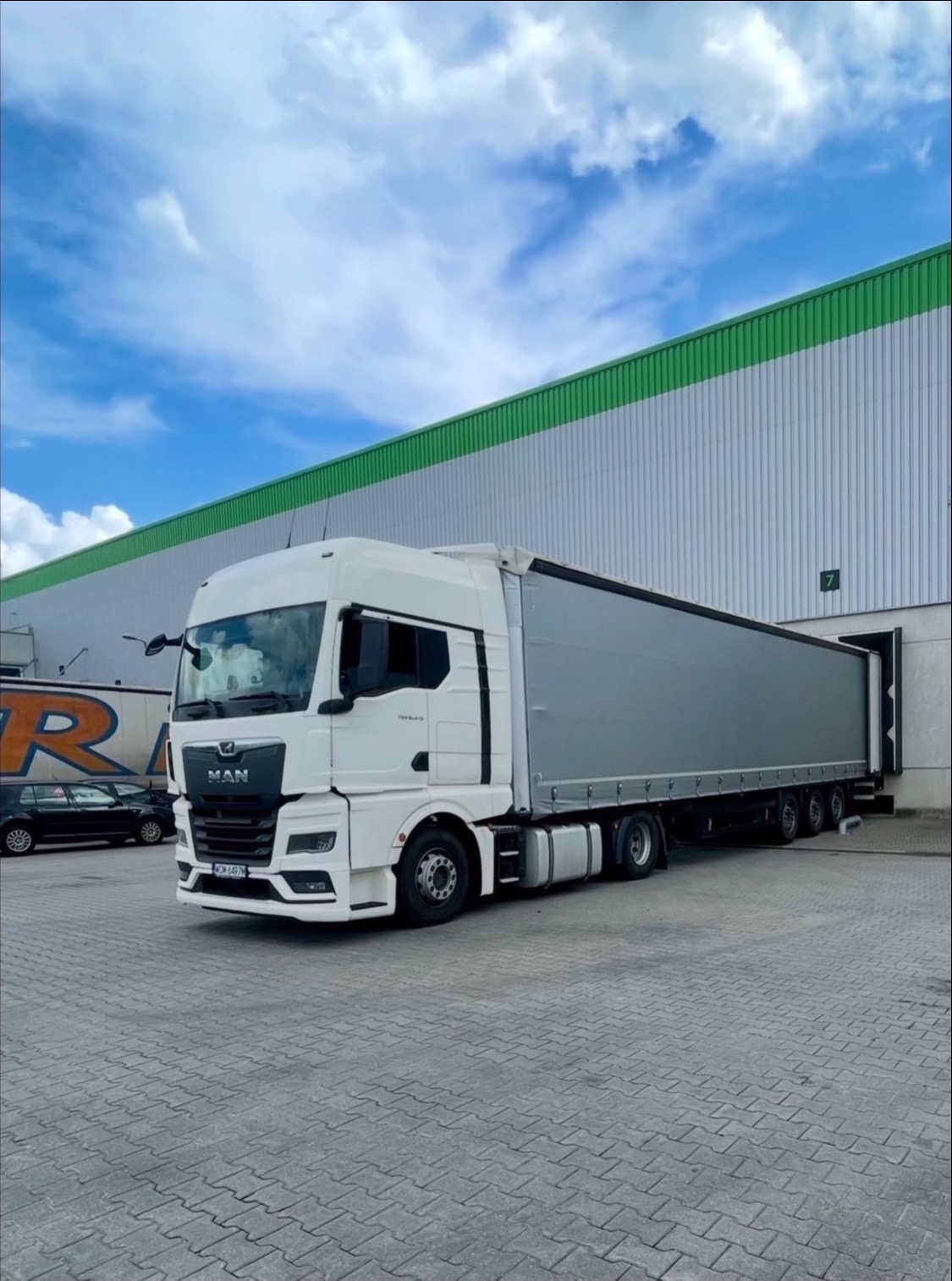 Biała ciężarówka MAN z naczepą na tle magazynu z zielonym dachem. Widoczne tablice rejestracyjne i inne pojazdy w tle. Dzień.