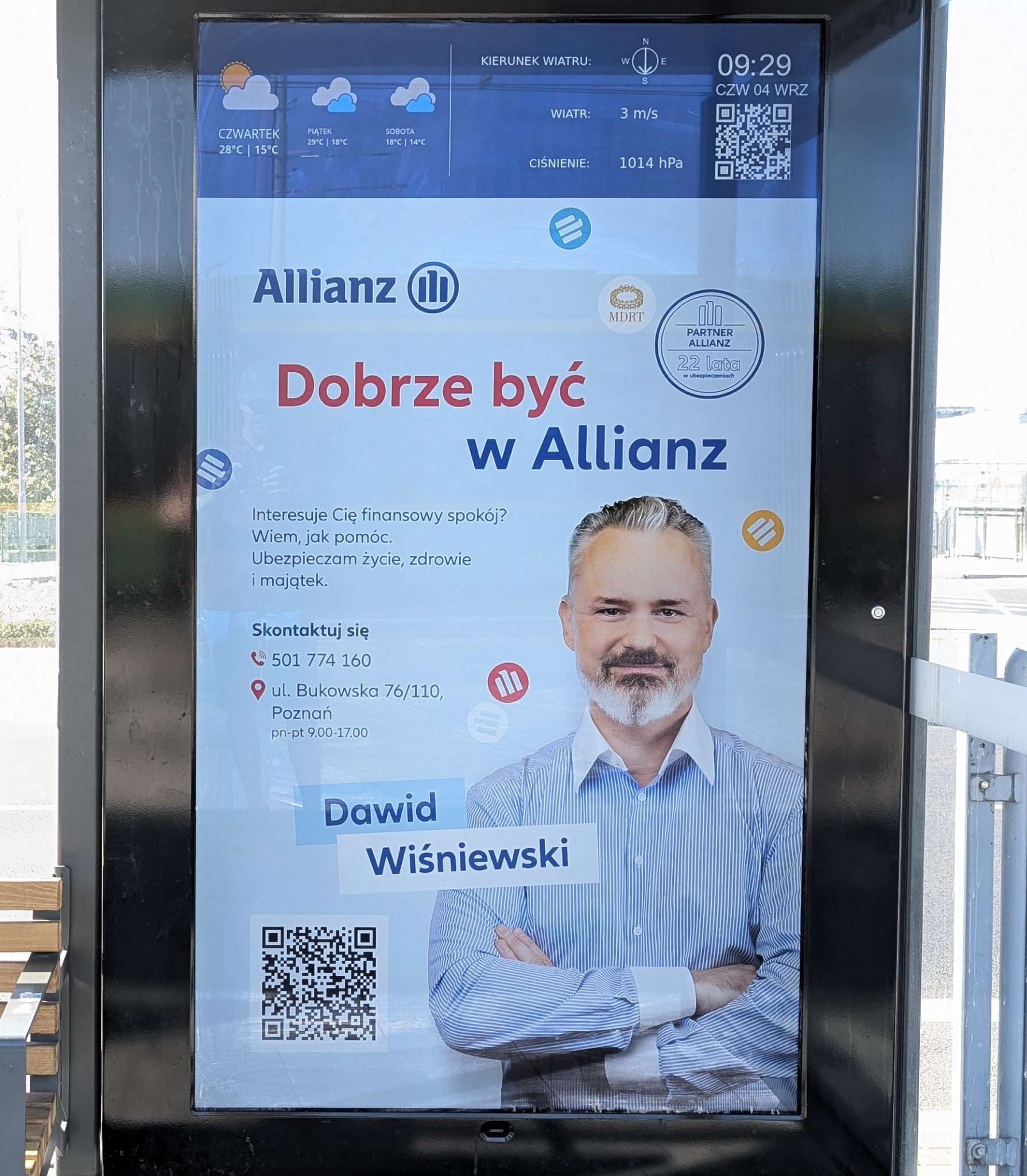 Reklama Allianz na przystanku autobusowym w Poznaniu. Widoczny agent ubezpieczeniowy Dawid Wiśniewski, dane kontaktowe i informacje o pogodzie. QR kod do zeskanowania.