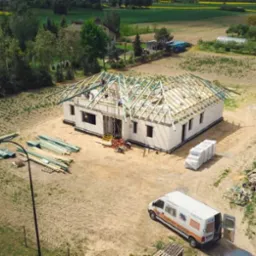 Budowa domu jednorodzinnego z białych bloczków, widok z góry. Częściowo zadaszona konstrukcja, na działce leżą materiały budowlane, stoi bus firmy budowlanej.