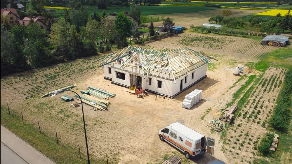 Budowa domu jednorodzinnego z białych bloczków, widok z góry. Częściowo zadaszona konstrukcja, na działce leżą materiały budowlane, stoi bus firmy budowlanej.