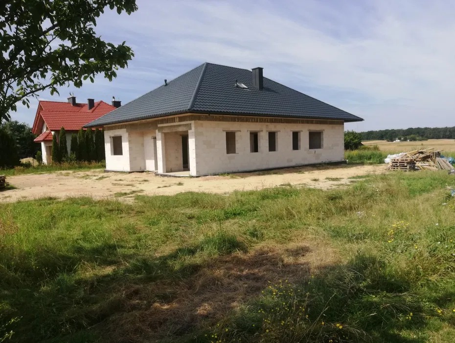 Dom w stanie surowym zamkniętym z ciemną dachówką, widok z zewnątrz na tle zieleni i pola. Budynek z pustaków z widocznymi otworami okiennymi.