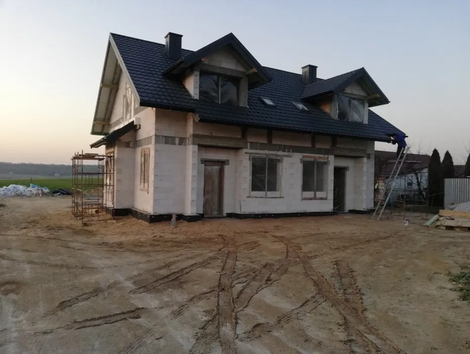 Dom jednorodzinny w trakcie budowy z czarnym dachem, widoczne rusztowania i osoba na drabinie, teren wokół domu przygotowany pod dalsze prace.