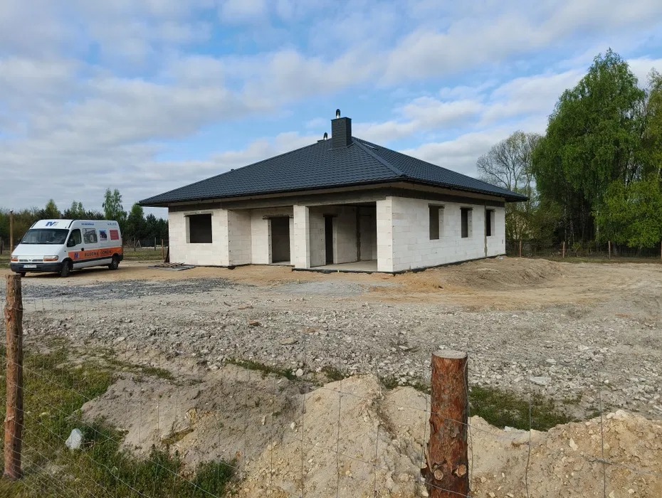 Surowy, parterowy dom z białego bloczka, z ciemnym dachem, w trakcie budowy. Na działce stoi biały bus. Widoczne drzewa w tle.