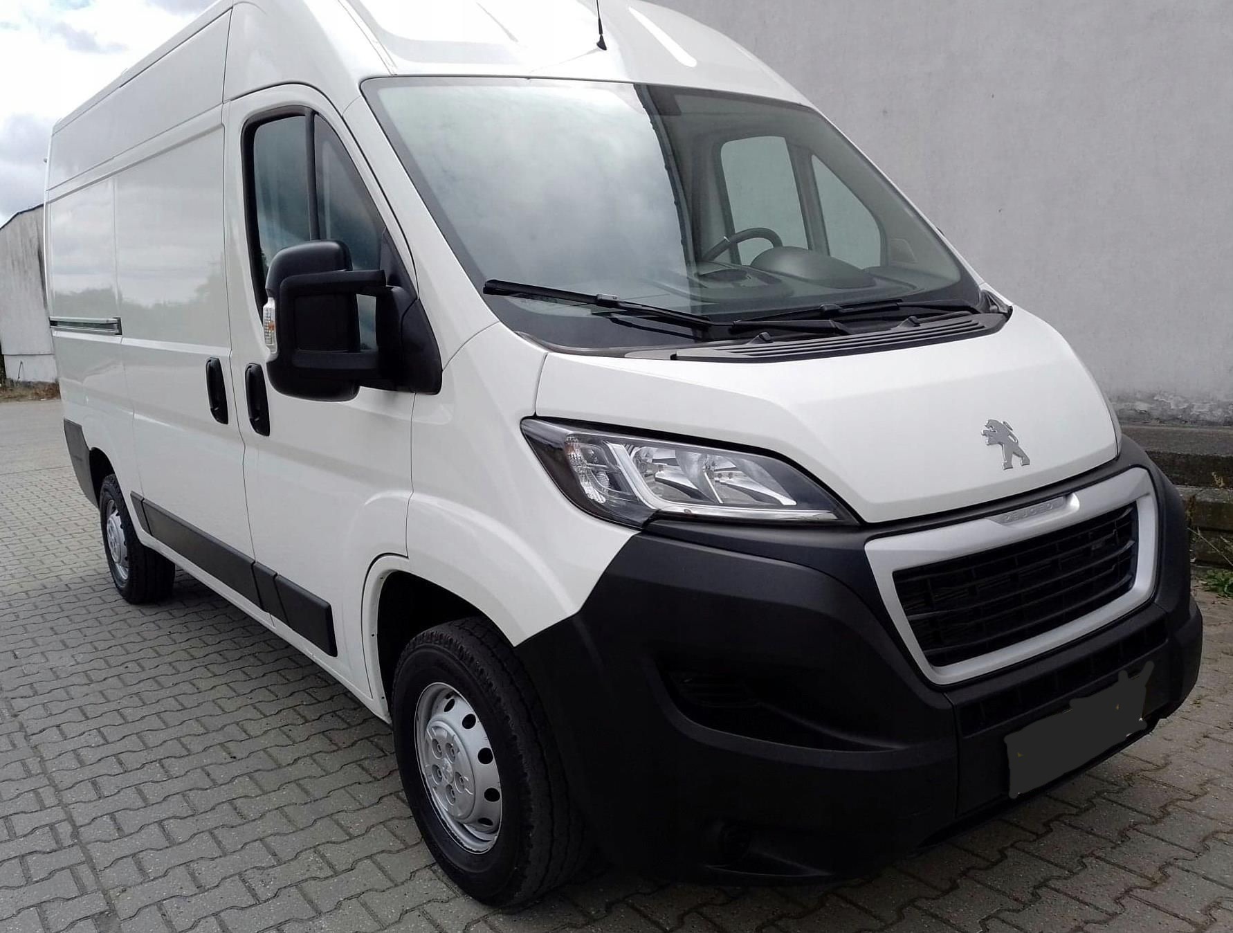 Biały bus Peugeot z widocznym logo na masce, zaparkowany na szarej kostce brukowej, prezentowany z perspektywy lekko z dołu.