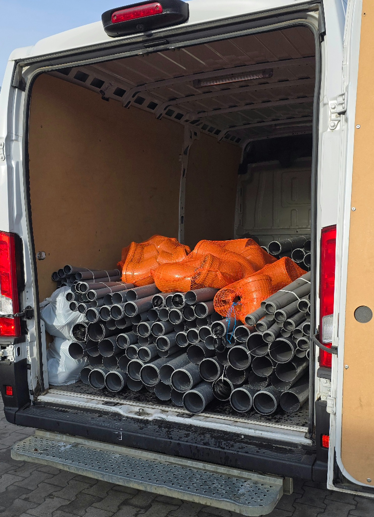 Wnętrze białego busa wypełnione szarymi rurami PCV i pomarańczowymi workami siatkowymi. Widoczne tylne drzwi i stopień wejściowy. Transport materiałów budowlanych.