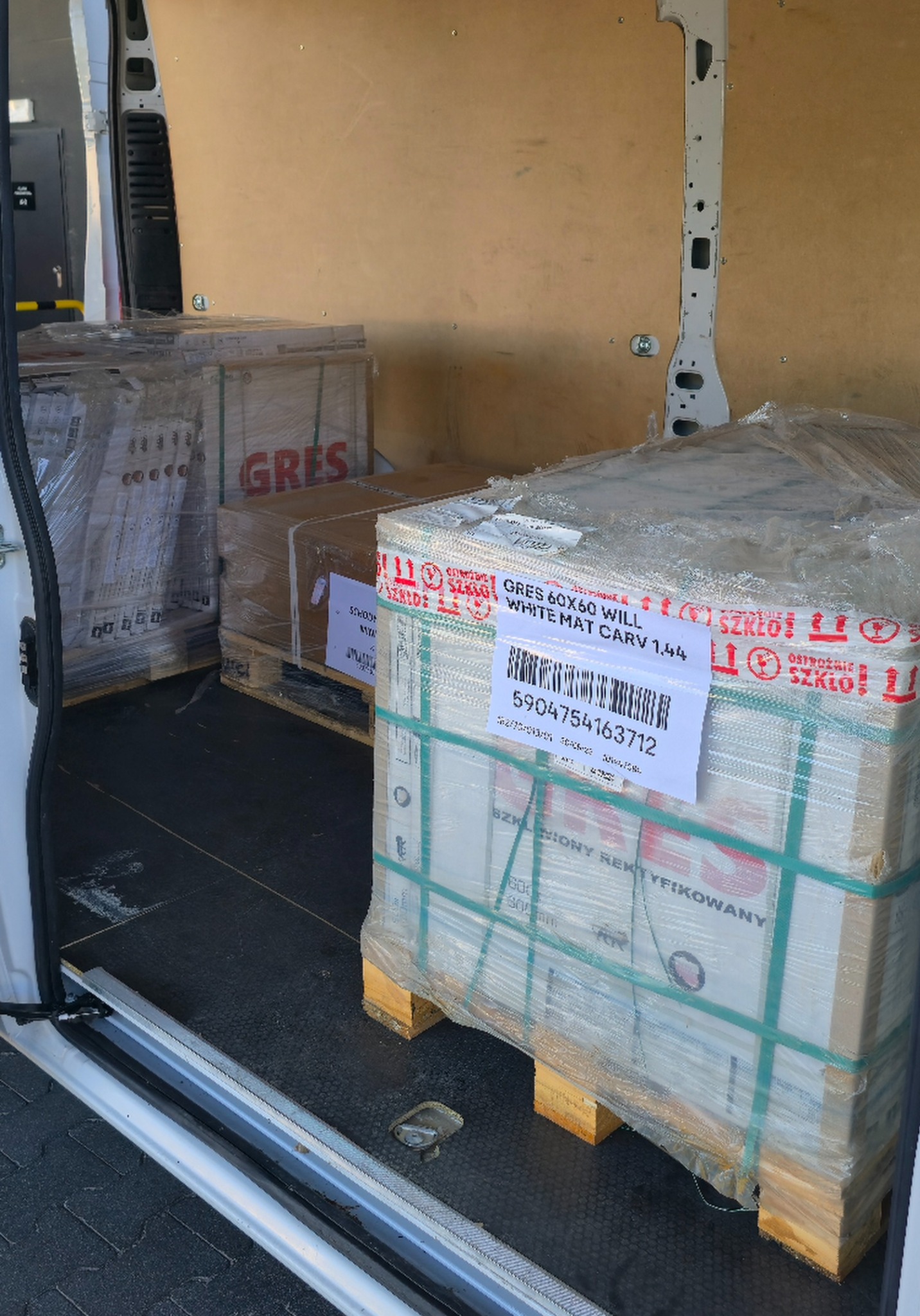 Paczki z płytkami GRES 60x60 WILL, zabezpieczone folią i umieszczone na paletach, wewnątrz przestrzeni ładunkowej busa. Widoczne etykiety z kodami kreskowymi.