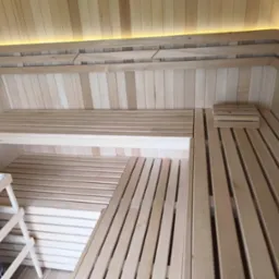 Jasne drewno, z którego wykonano wnętrze sauny. Widoczne ławki i oparcia, a także piec z kamieniami. Oświetlenie LED podkreśla naturalny charakter drewna.