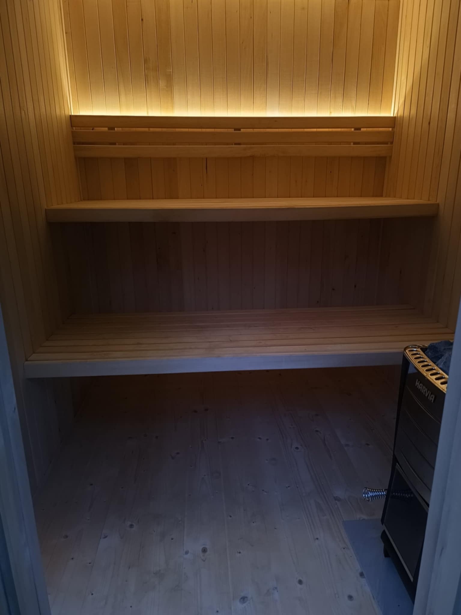 Wnętrze sauny z drewnianymi ławkami i piecem Harvia w rogu, oświetlone subtelnym światłem. Widok od wejścia.