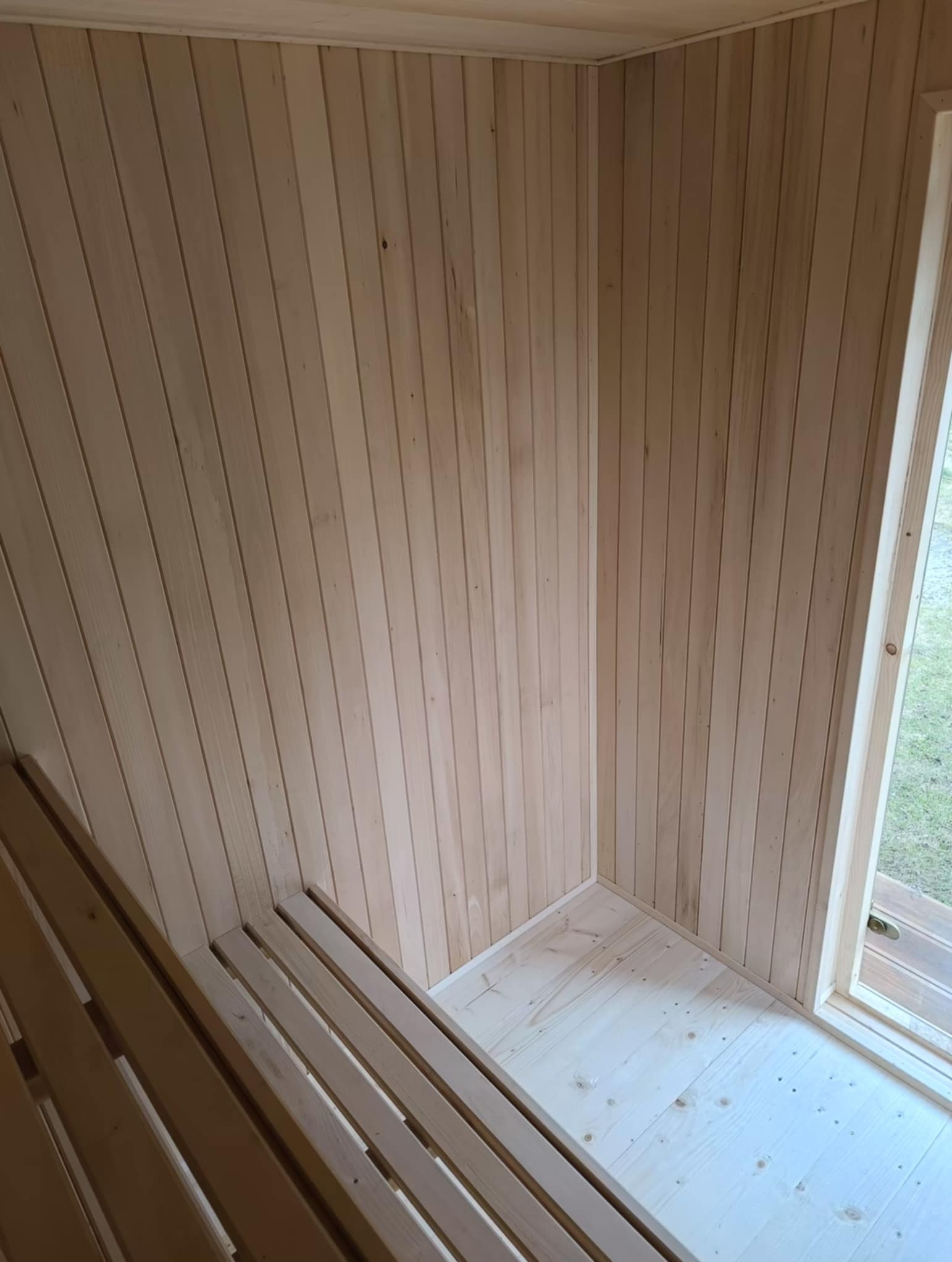 Jasne drewno w saunie: deski na ścianach, podłodze i ławki. Widoczne słoje drewna i okno z widokiem na zewnątrz. Minimalistyczny design.