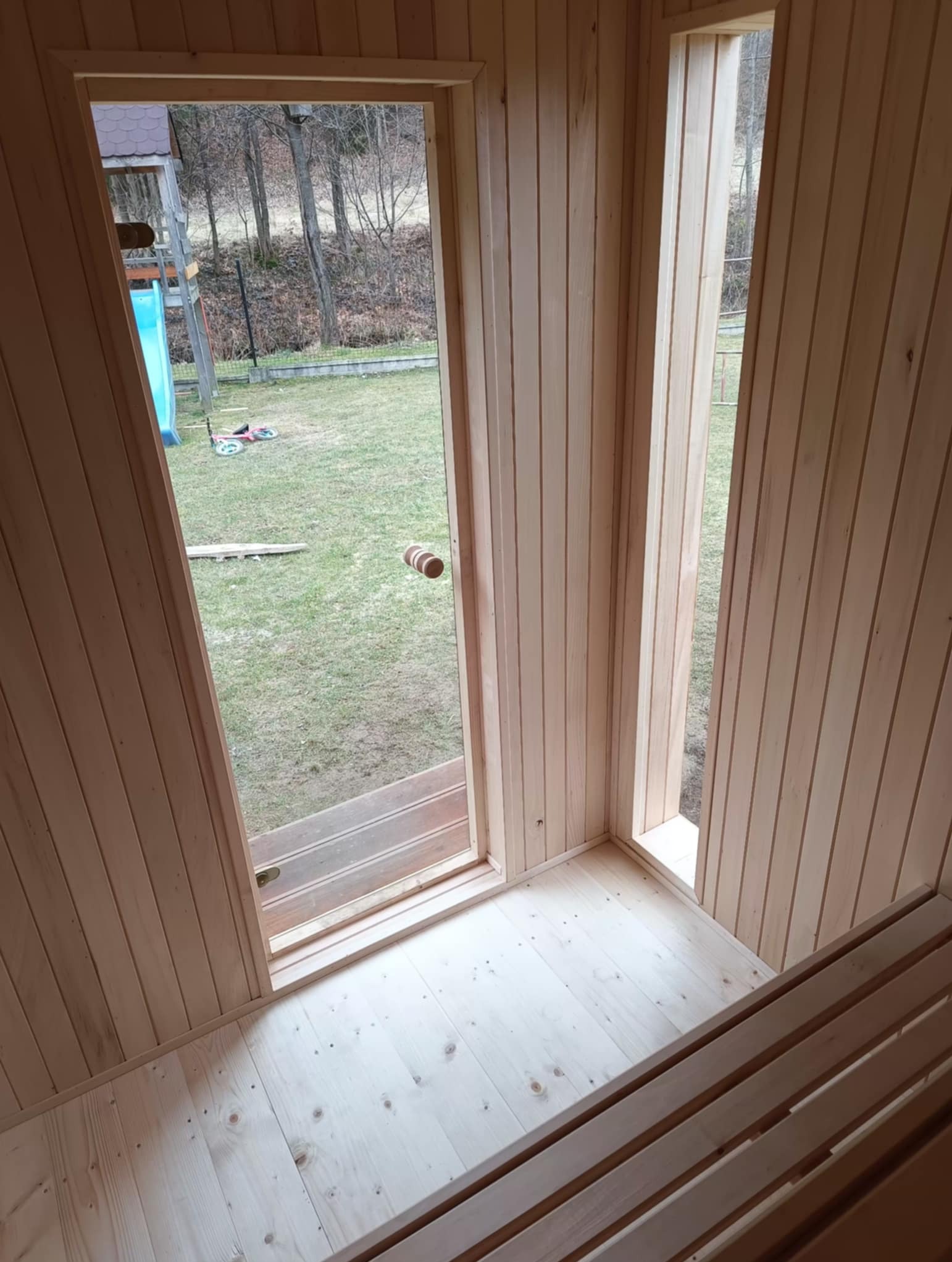 Wnętrze nowej sauny z boazerią i drewnianą ławką, widok na zewnątrz przez otwarte drzwi i okno. Widoczny trawnik i plac zabaw.