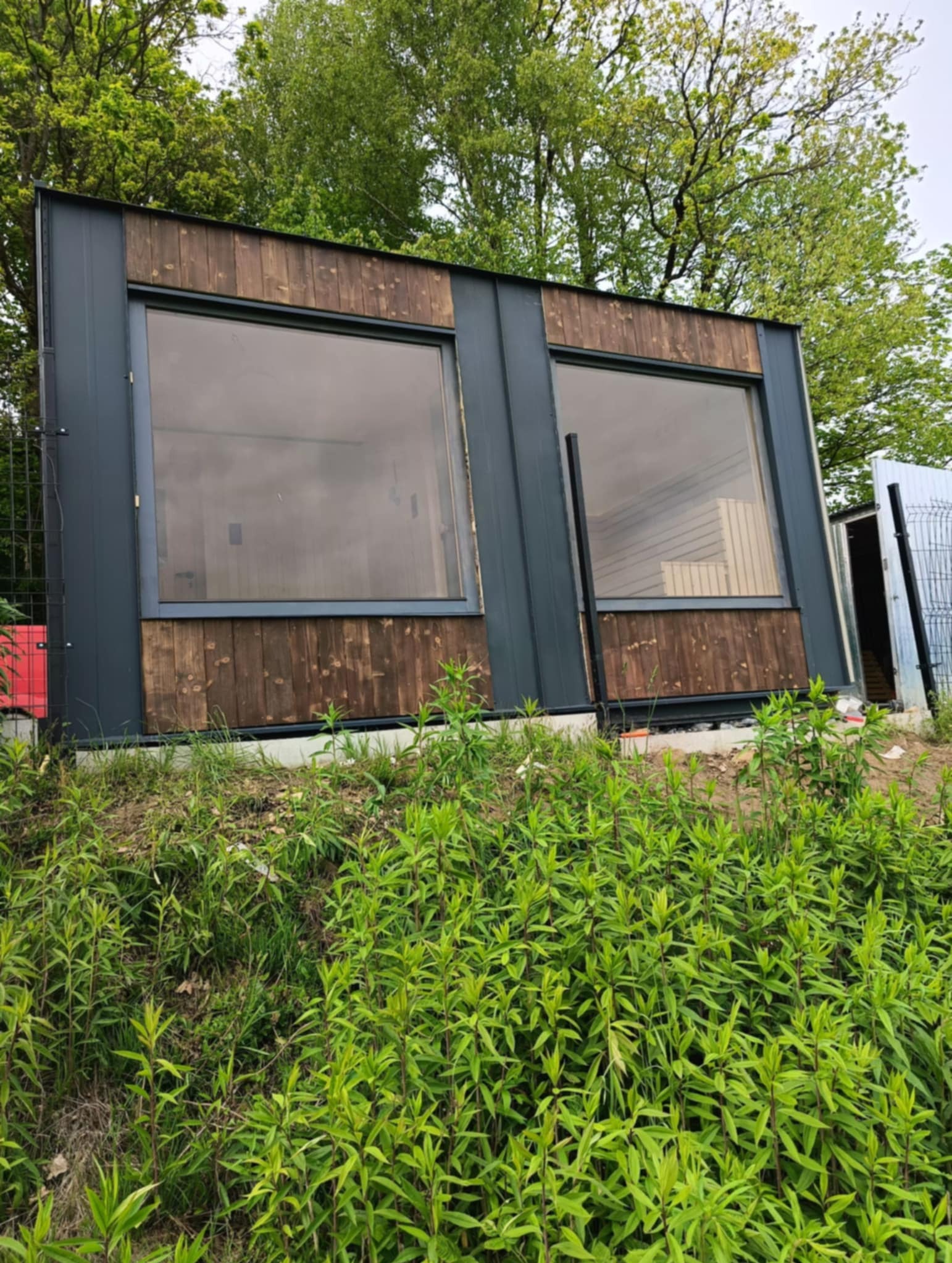 Nowoczesna sauna zewnętrzna z drewnianym wykończeniem i dużymi oknami, otoczona zielenią. Widoczne elementy konstrukcyjne i częściowo wnętrze.