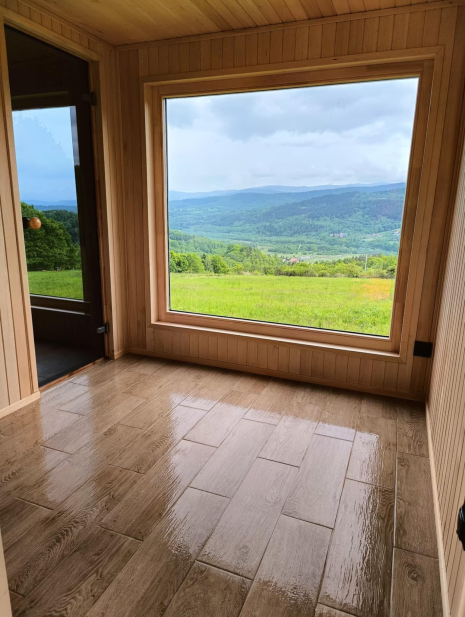 Wnętrze drewnianej sauny z dużym oknem panoramicznym z widokiem na zalesione wzgórza. Podłoga wyłożona płytkami imitującymi drewno.