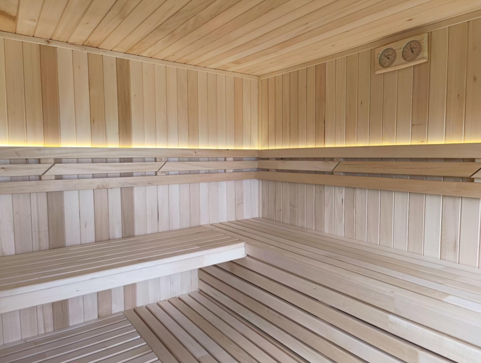 Jasne drewno w saunie z termometrami i podświetleniem LED. Widoczne ławki i boazeria, tworzące ciepłą atmosferę. Drewniane wykończenie.