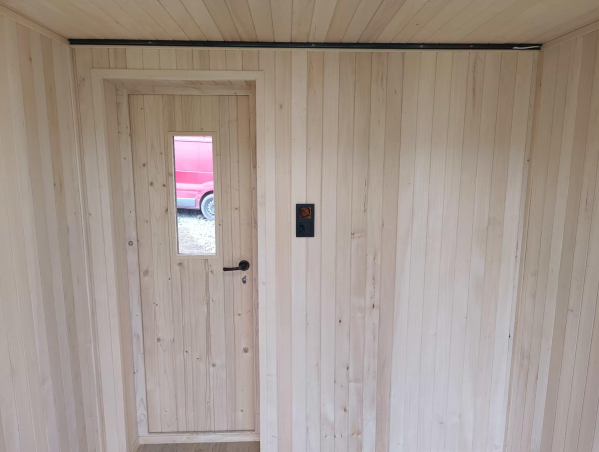Wnętrze sauny z jasnego drewna: drzwi z małym okienkiem, czarny włącznik na ścianie. Widoczny fragment czerwonego samochodu przez okno.