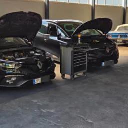 Dwa czarne Renault z otwartymi maskami w warsztacie samochodowym, w tle narzędziowy wózek i niebieski klasyk. Warsztat oświetlony przez duże okna.