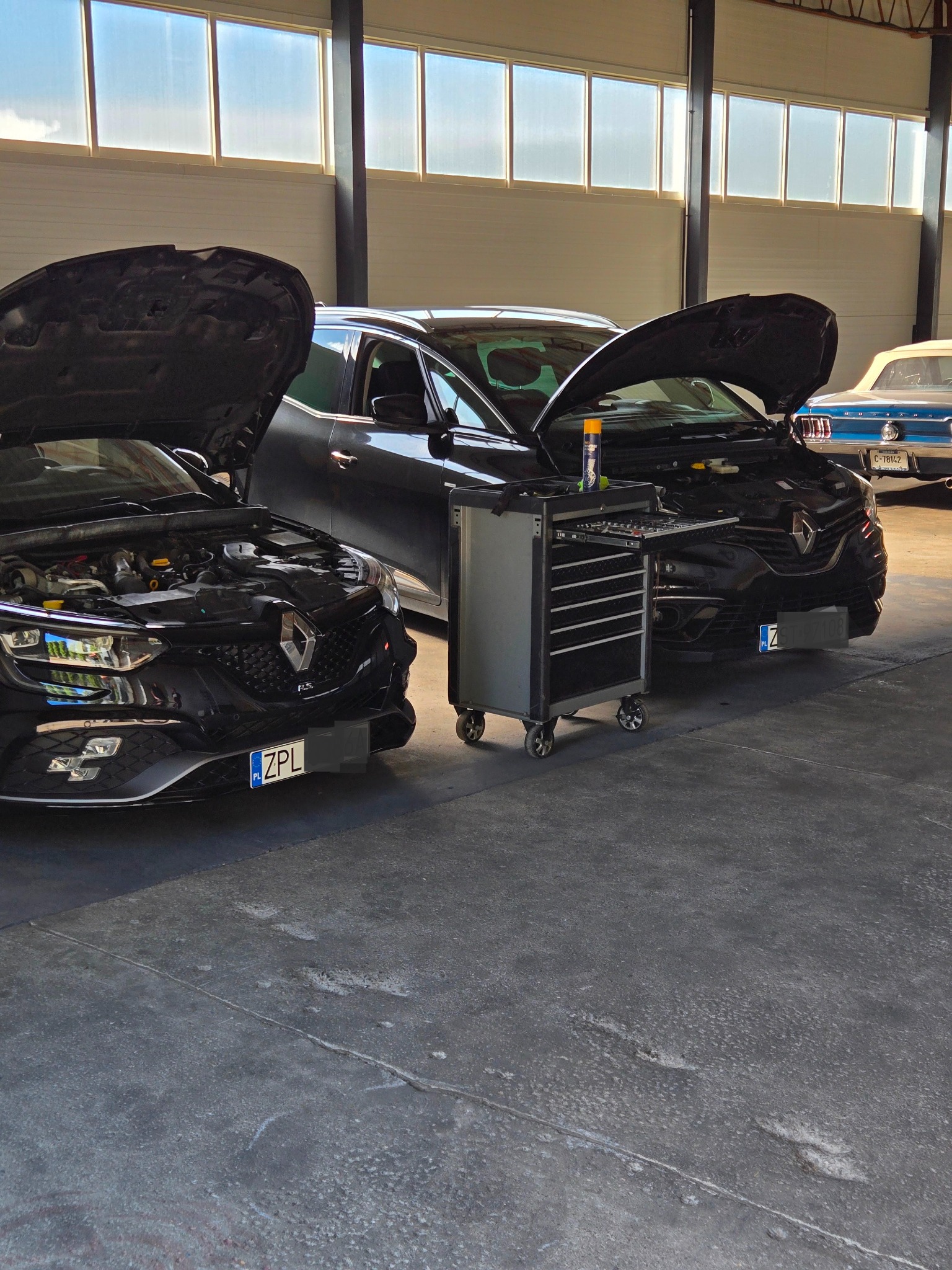 Dwa czarne Renault z otwartymi maskami w warsztacie samochodowym, w tle narzędziowy wózek i niebieski klasyk. Warsztat oświetlony przez duże okna.