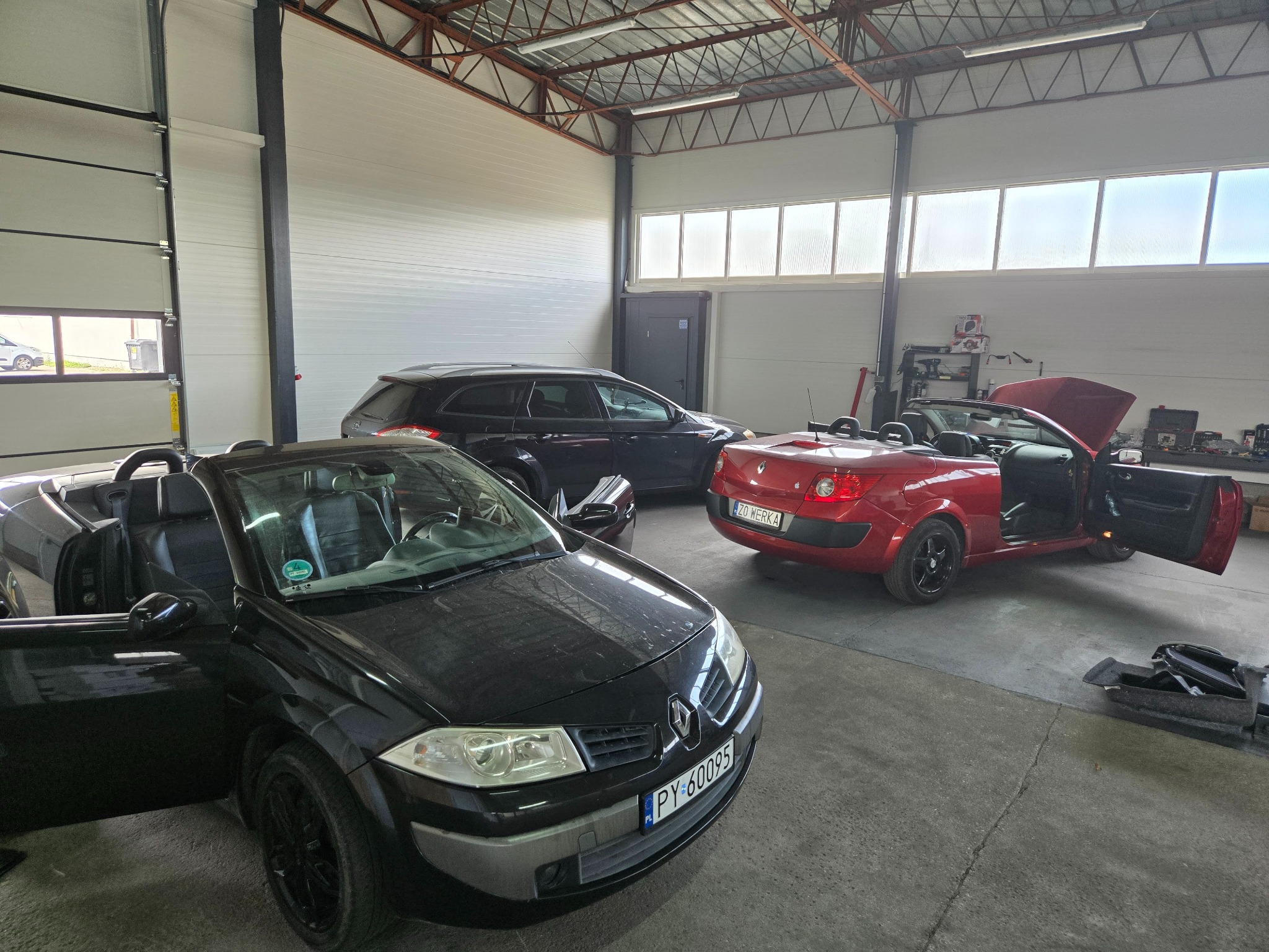 Warsztat z trzema autami: czarnym i czerwonym kabrioletem Renault Megane z otwartymi drzwiami oraz ciemnym kombi w tle. Widoczne wyposażenie warsztatu.