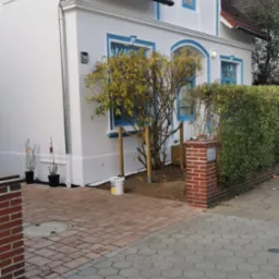 Biały dom z niebieskimi akcentami po renowacji, widoczny z zewnątrz, z odświeżoną elewacją i nowymi panelami słonecznymi na dachu. Na pierwszym planie wiadro z farbą.