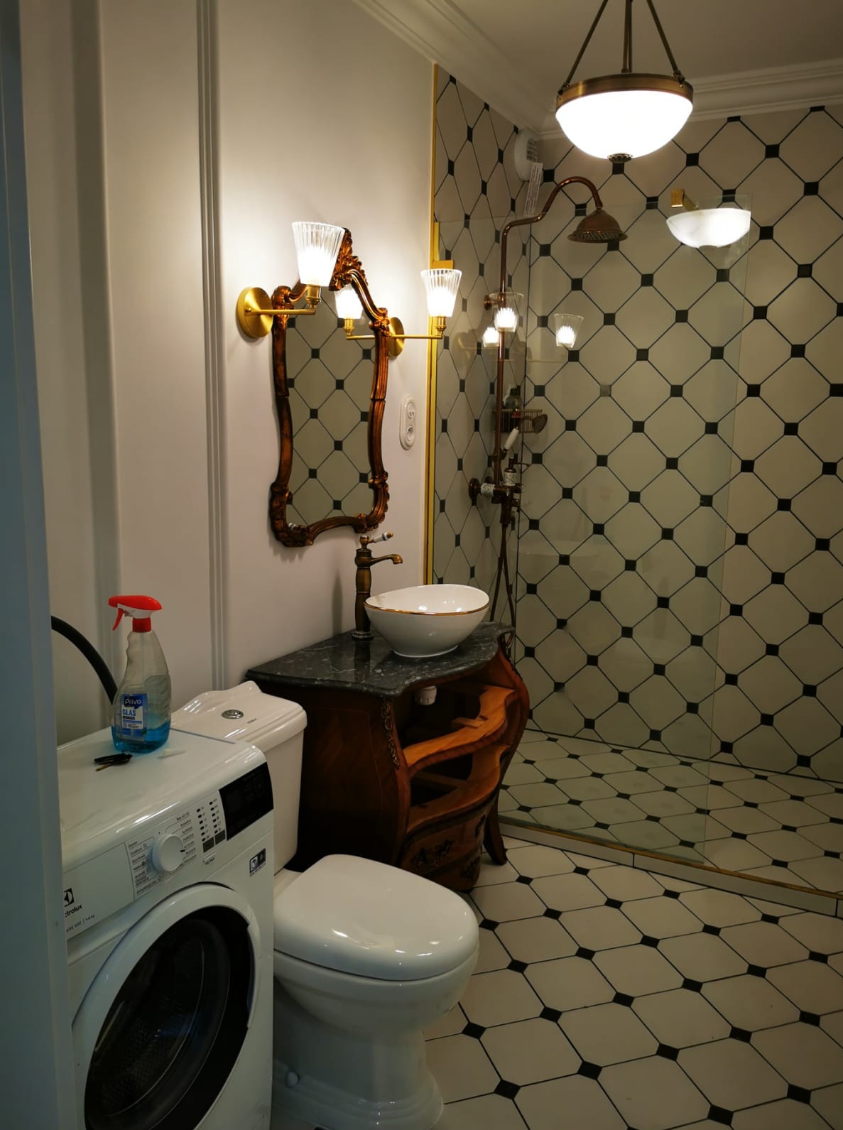 Elegancka łazienka z umywalką na zdobionej szafce, lustrem w złotej ramie i kabiną prysznicową w stylu retro, z pralką po lewej stronie.