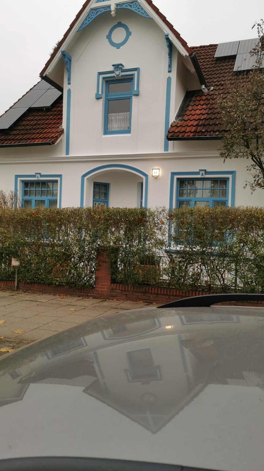 Biały dom z niebieskimi akcentami, widoczny zza żywopłotu i odbijający się w karoserii samochodu. Na dachu panele słoneczne.