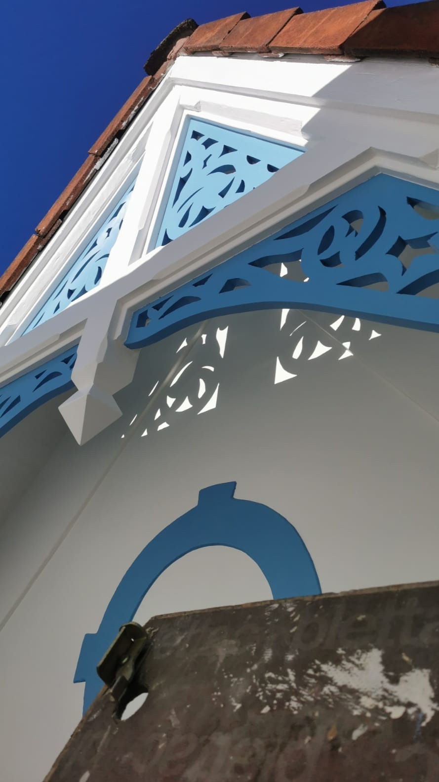Detal elewacji budynku po renowacji, z dekoracyjnymi, ażurowymi elementami w kolorze błękitnym i białym, widzianymi pod kątem, z fragmentem dachu z dachówką.