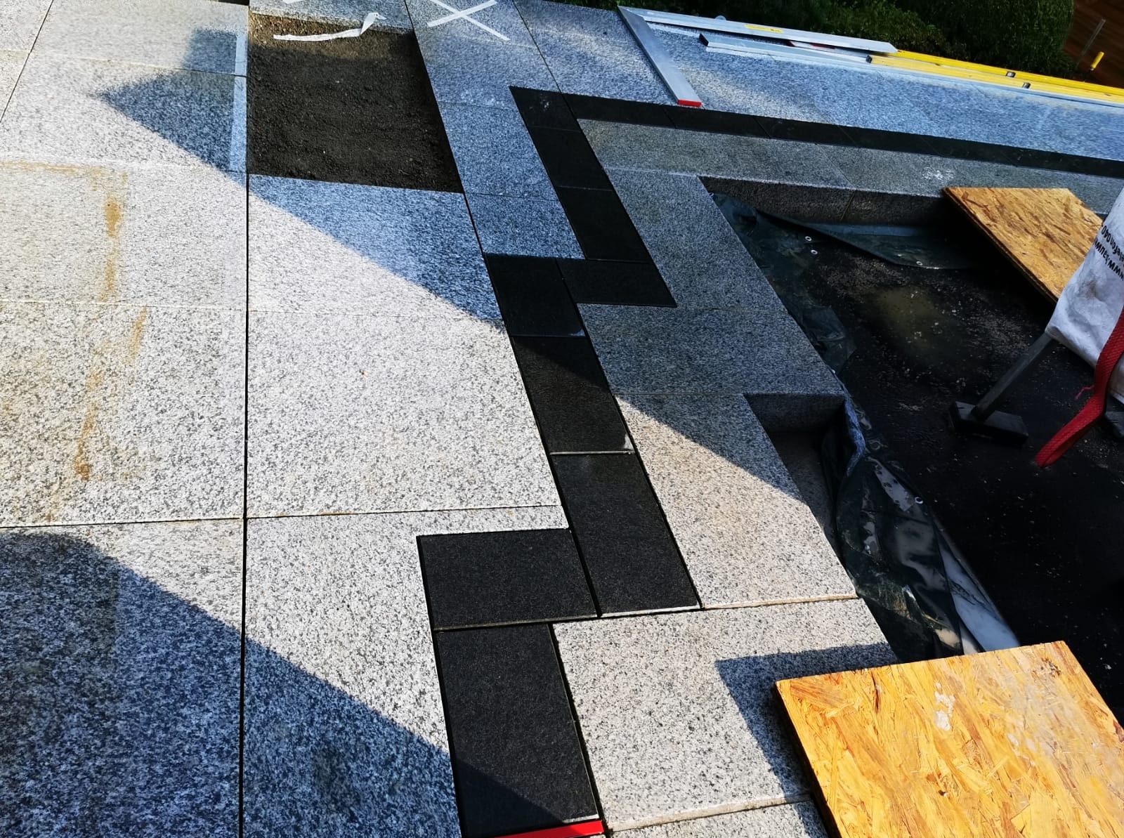 Układanie szarych i czarnych płyt granitowych na zewnątrz, widoczne fragmenty podłoża i poziomica, praca w toku.