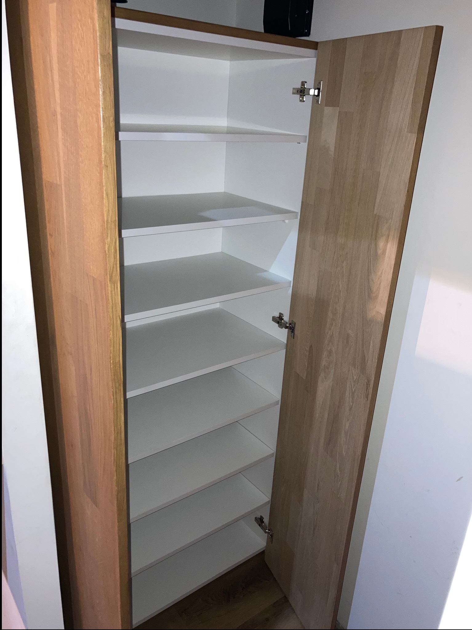 Otwarta szafa wnękowa z półkami, białe wnętrze, drewniane drzwi z widocznymi zawiasami, minimalistyczny design, oświetlenie od góry.