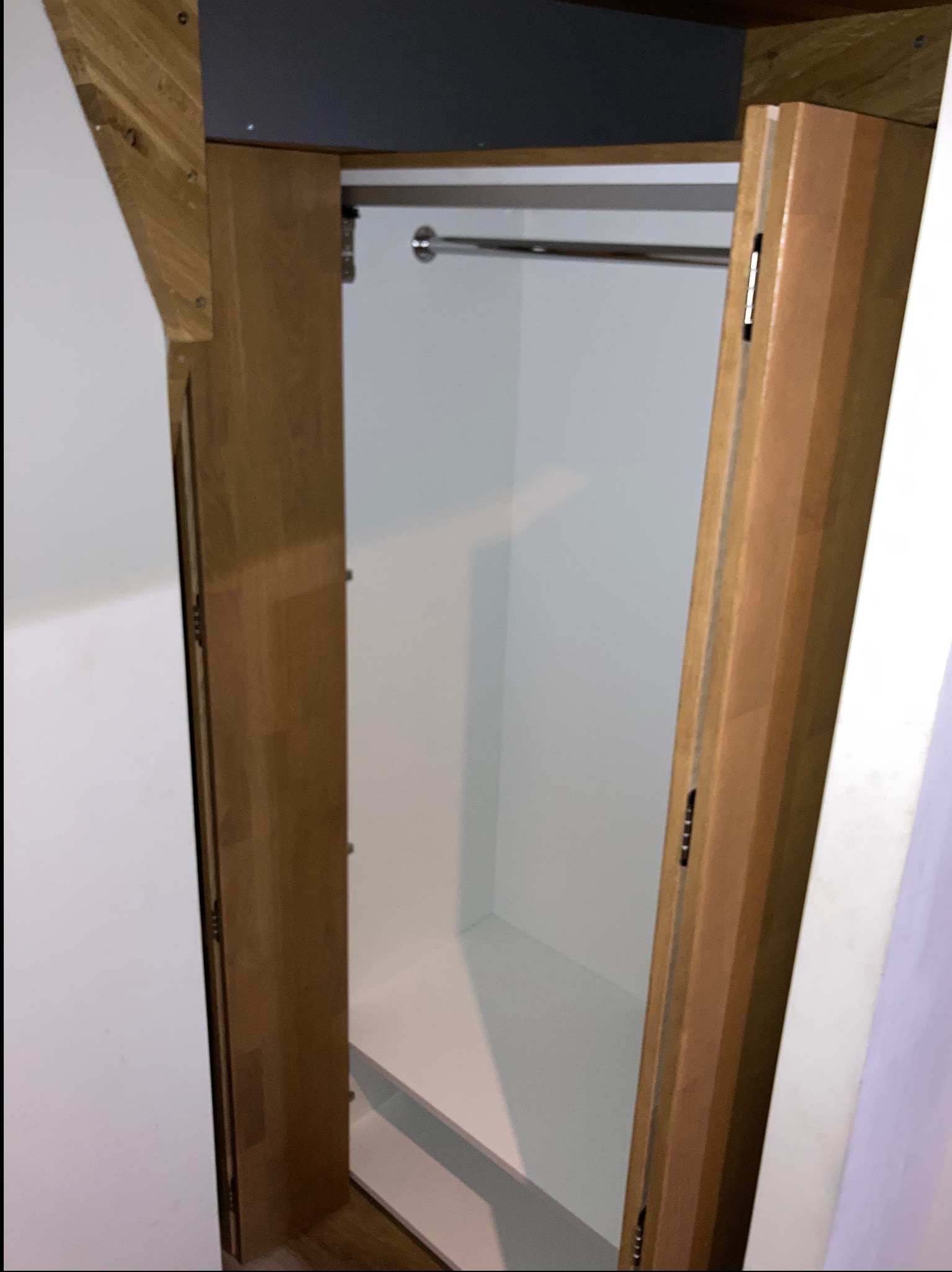 Otwarta szafa wnękowa z drewnianymi drzwiami i metalowym drążkiem na ubrania, widoczna z perspektywy wnętrza, z elementami konstrukcyjnymi na górze.