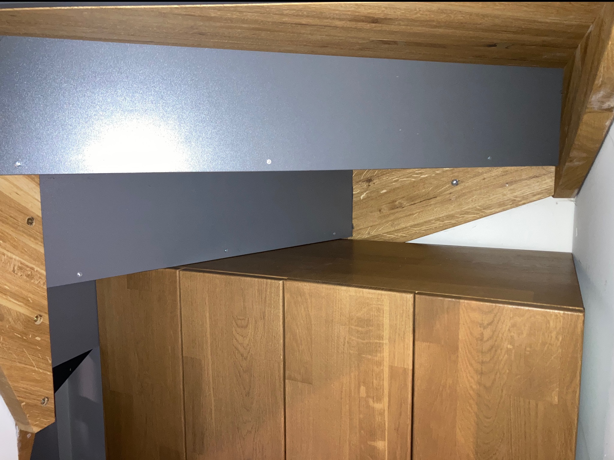 Drewniana szafka pod schodami z szarą płytą, widoczne elementy konstrukcyjne schodów z jasnego drewna. Nietypowe zagospodarowanie przestrzeni.