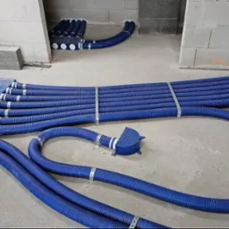 Niebieskie rury wentylacyjne ułożone na podłodze w surowym wnętrzu, z widocznymi elementami montażowymi i fragmentami konstrukcji budynku w tle.