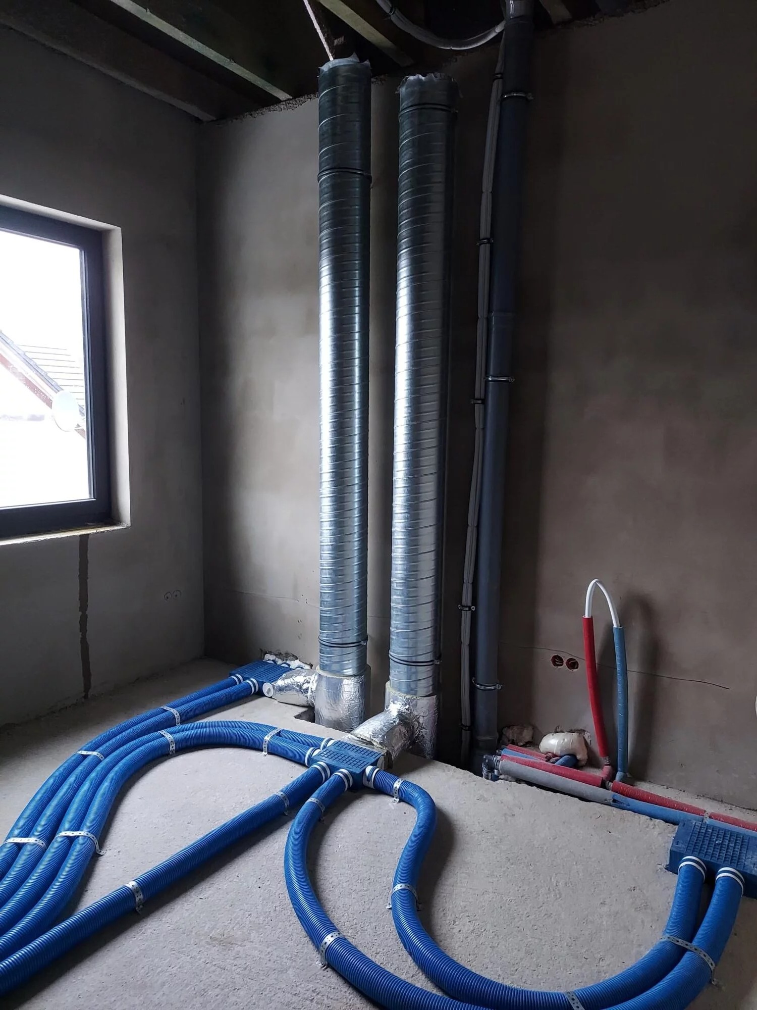 Surowy stan: instalacja wentylacji z rurami w kolorze niebieskim i pionowymi, metalowymi przewodami w nowym budynku. Widoczne okno i surowy beton.