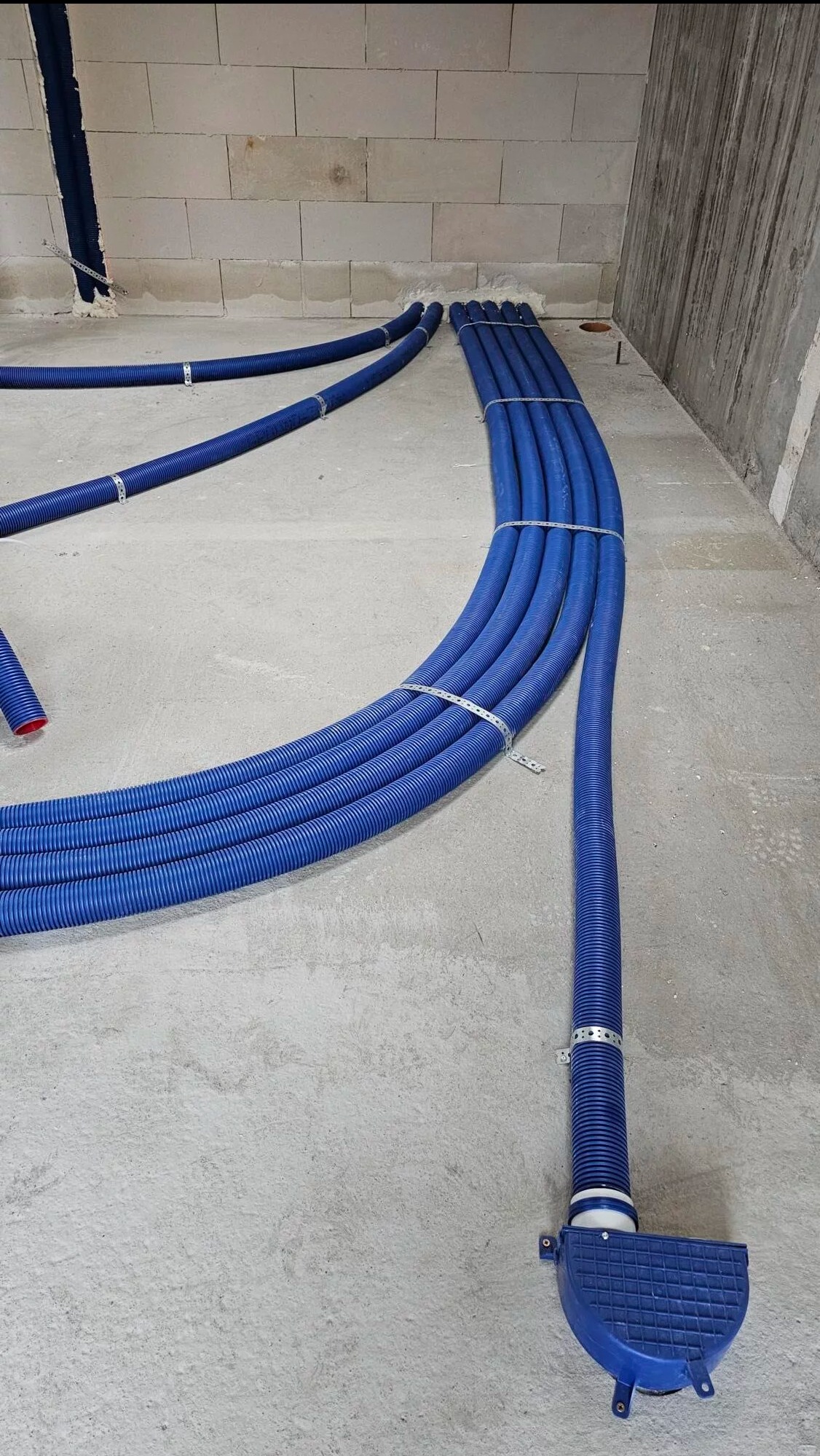 Niebieskie rury wentylacyjne, częściowo zamocowane do ściany, ułożone na szarym betonie. Widoczny fragment ściany z betonu komórkowego.