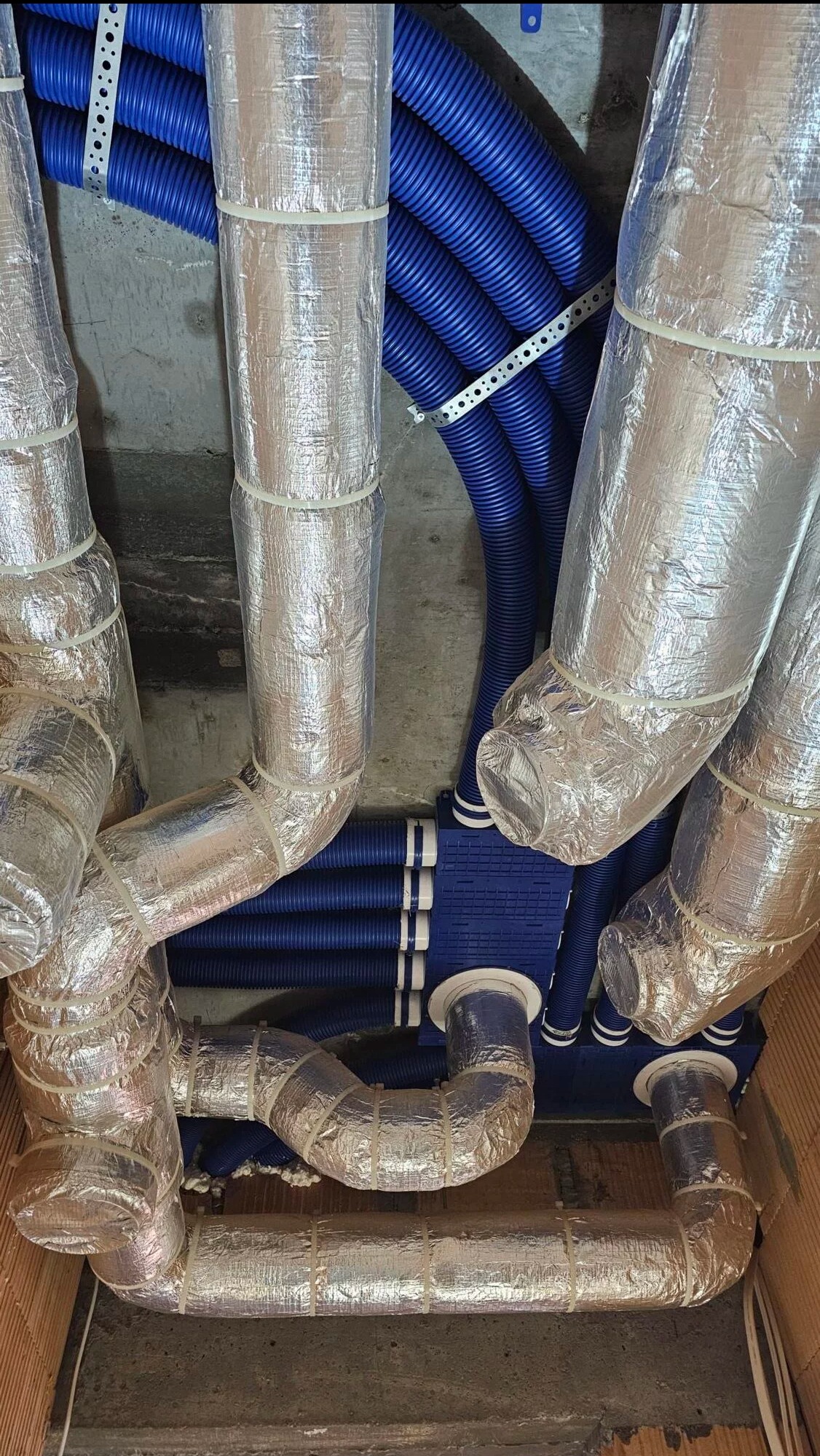 Niebieskie rury wentylacyjne z folią izolacyjną, połączone z rozdzielaczem, zamontowane pod sufitem. Widok z dołu ukazuje skomplikowany system wentylacji.