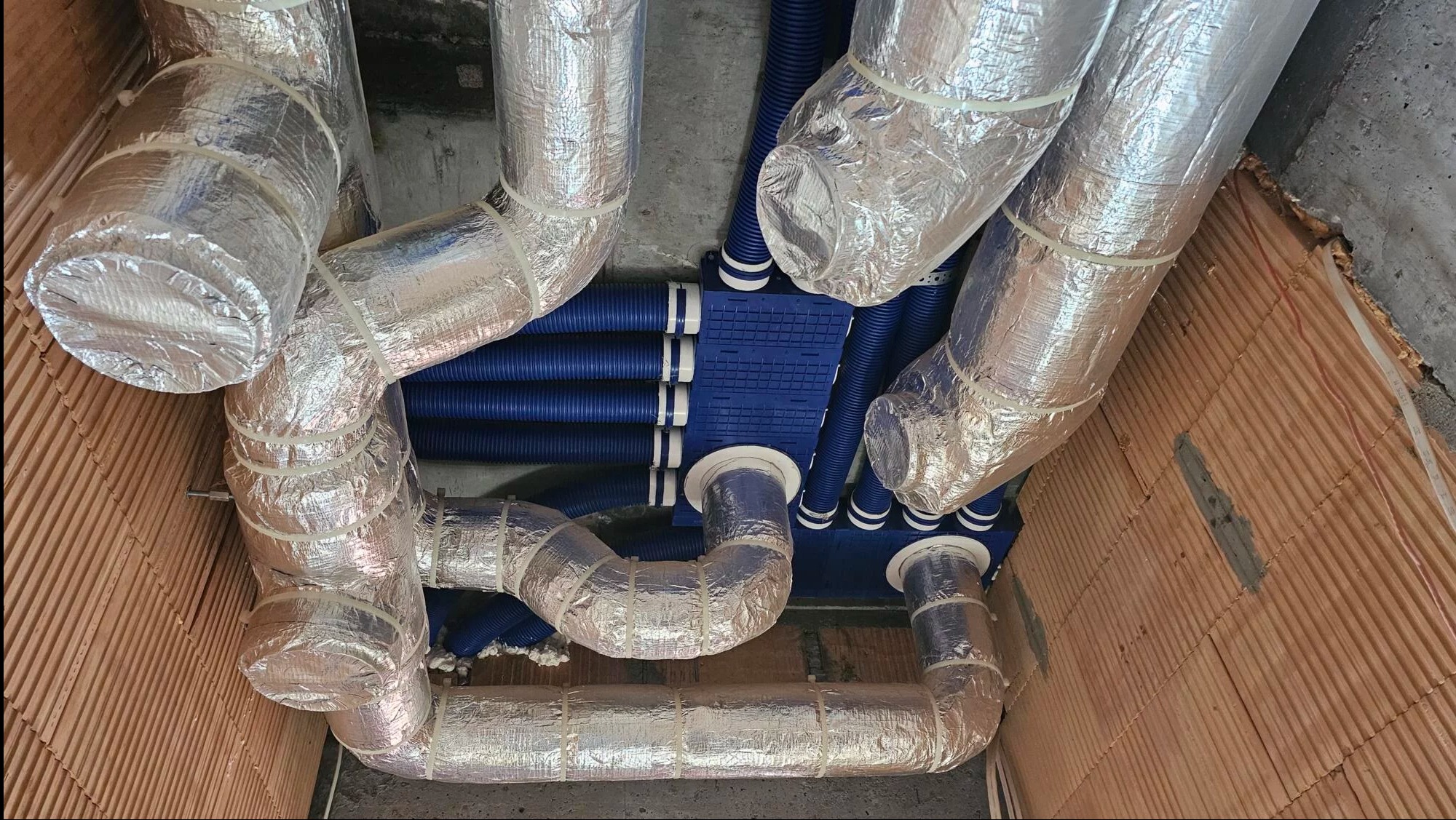 Rozbudowana instalacja wentylacyjna z niebieskimi rurami i izolowanymi przewodami aluminiowymi, zamontowana pod sufitem, widoczna z perspektywy pod kątem.
