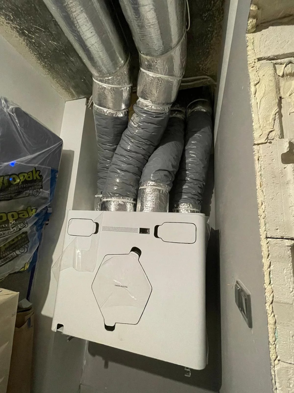 Biała jednostka rekuperacyjna z szarymi, elastycznymi przewodami wentylacyjnymi w pomieszczeniu z surowymi ścianami. Widoczne opakowania po elementach instalacji.