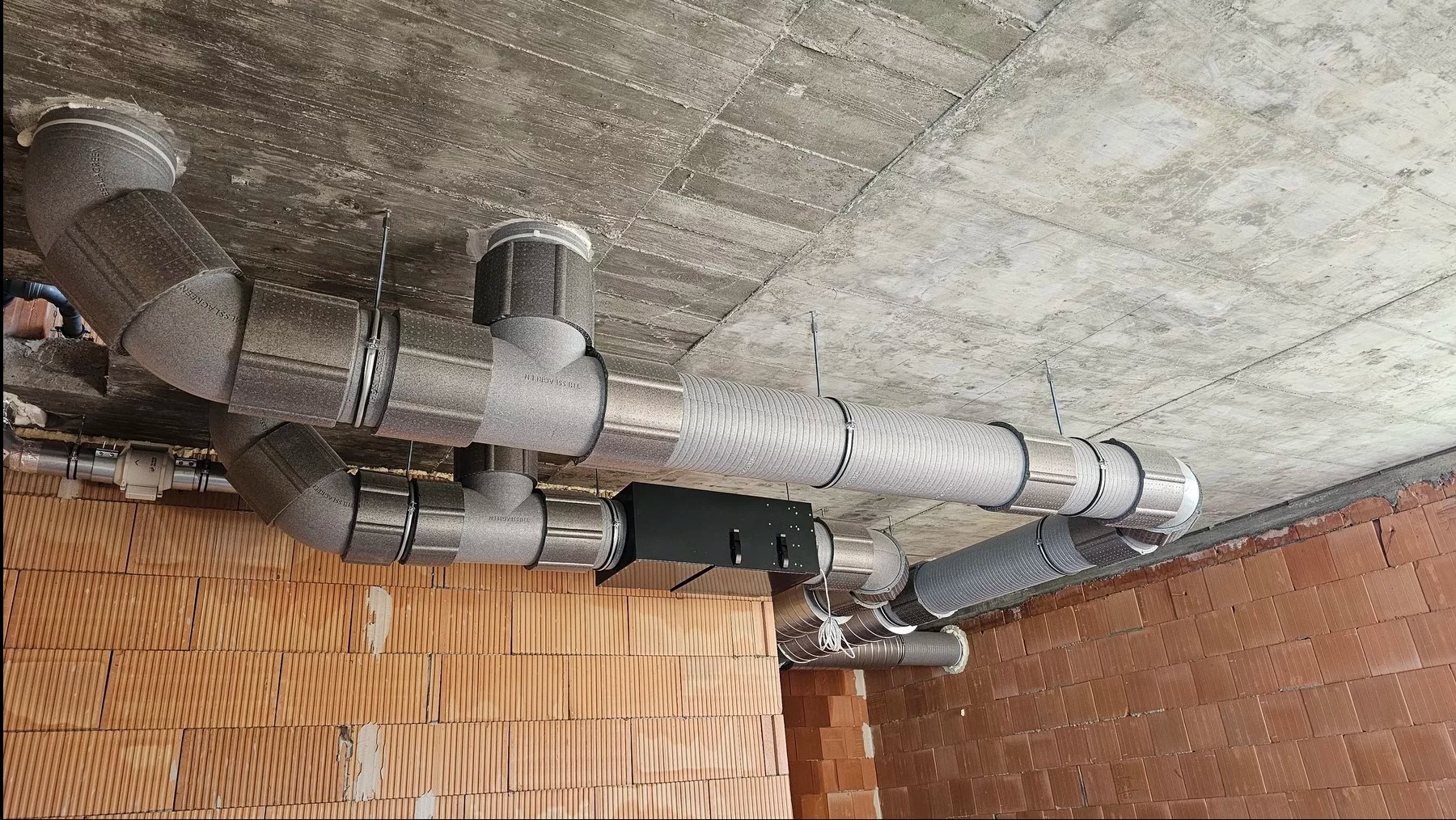 Kompleksowa instalacja wentylacyjna z szarych rur na surowym suficie betonowym i ścianie z czerwonej cegły. Widoczne złączenia i elementy montażowe.