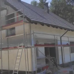 Dom w trakcie ocieplania elewacji, widoczne rusztowania, częściowo ocieplona ściana, styropian i narzędzia. Tło stanowią drzewa.
