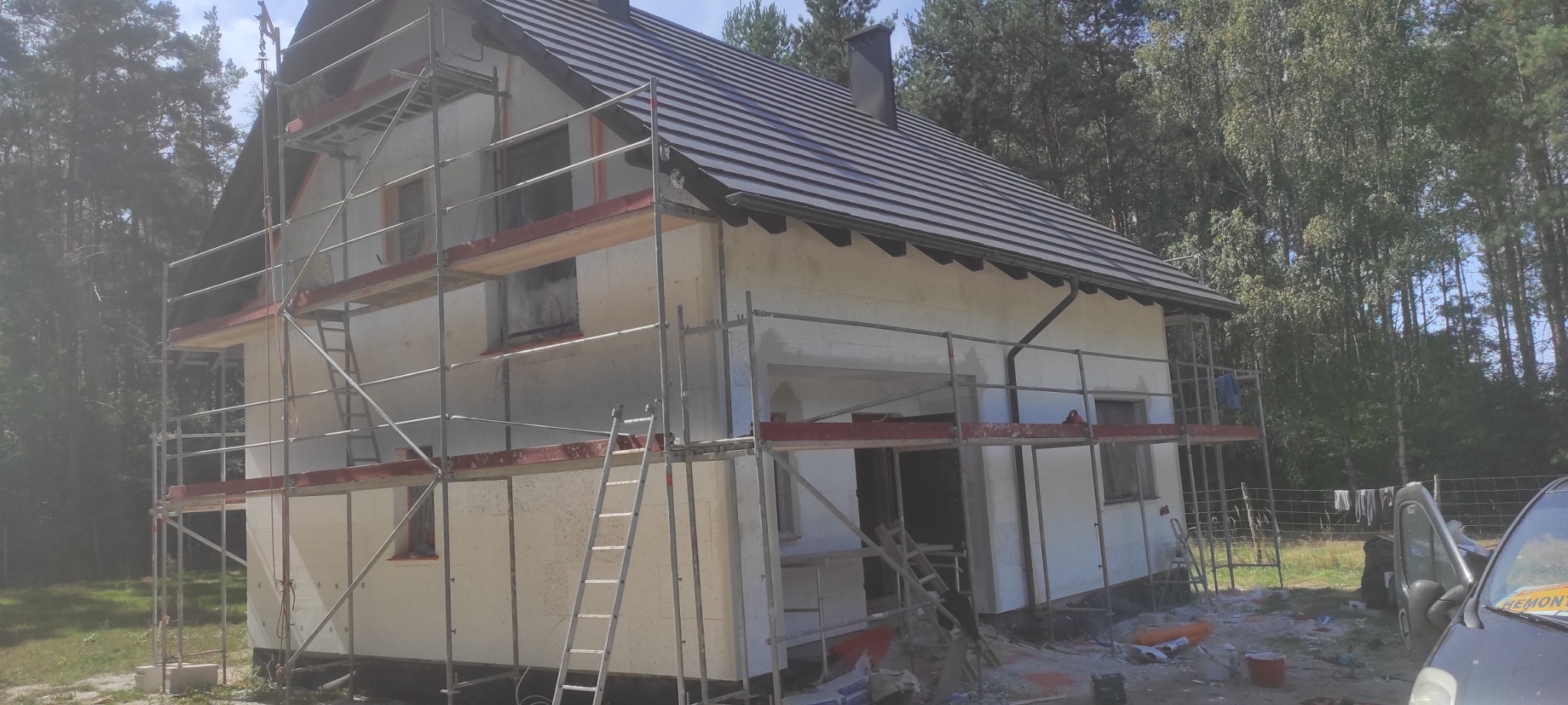 Dom w trakcie ocieplania elewacji, widoczne rusztowania, częściowo ocieplona ściana, styropian i narzędzia. Tło stanowią drzewa.