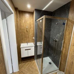 Jakub Jasiński Interior Bud - Wykończona łazienka z kabiną prysznicową, umywalką z szafką i drewnopodobnymi płytkami. Nowoczesny design z akcentami drewna i szarości.