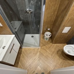 Jakub Jasiński Interior Bud - Wykończona łazienka z kabiną prysznicową, umywalką z szafką i toaletą. Uwagę przykuwają płytki imitujące drewno ułożone w jodełkę na podłodze i ścianach.