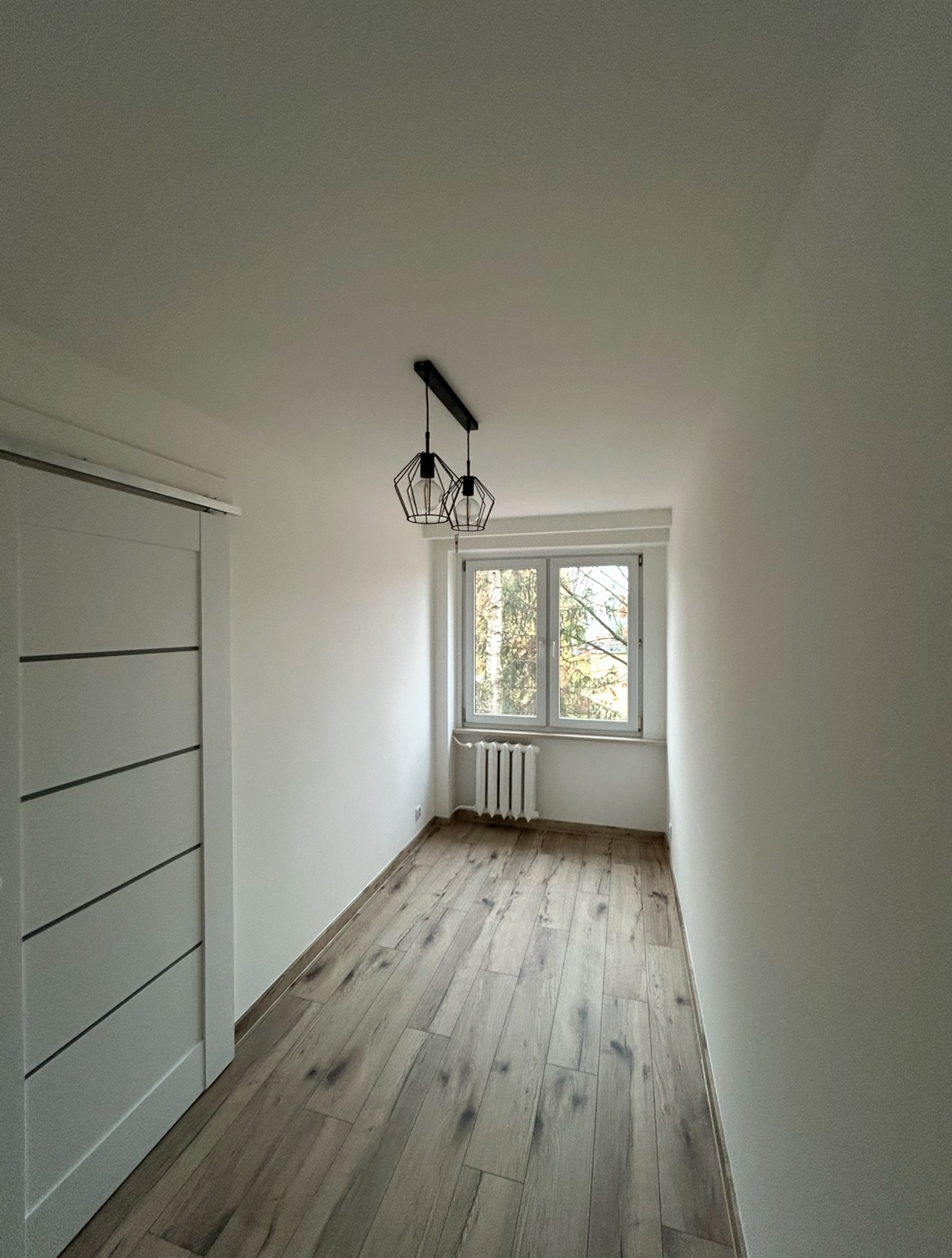 Jasne panele podłogowe ułożone w pokoju po remoncie w Stalowej Woli. Ściany pomalowane na biało, widoczne okno z grzejnikiem i nowoczesna lampa.