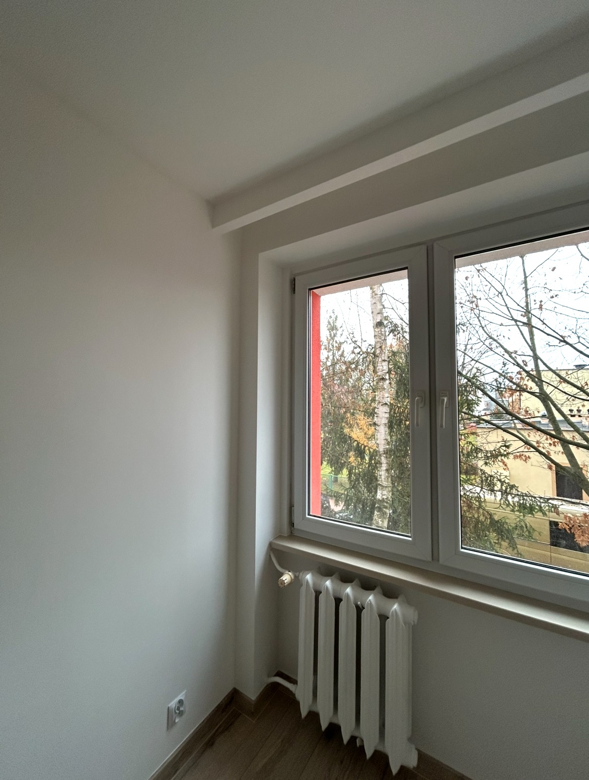 Jasne, odświeżone wnętrze pokoju po malowaniu, z białymi ścianami, grzejnikiem i oknem z widokiem na drzewa. Jeden element ramy okiennej jest pomalowany na czerwono.