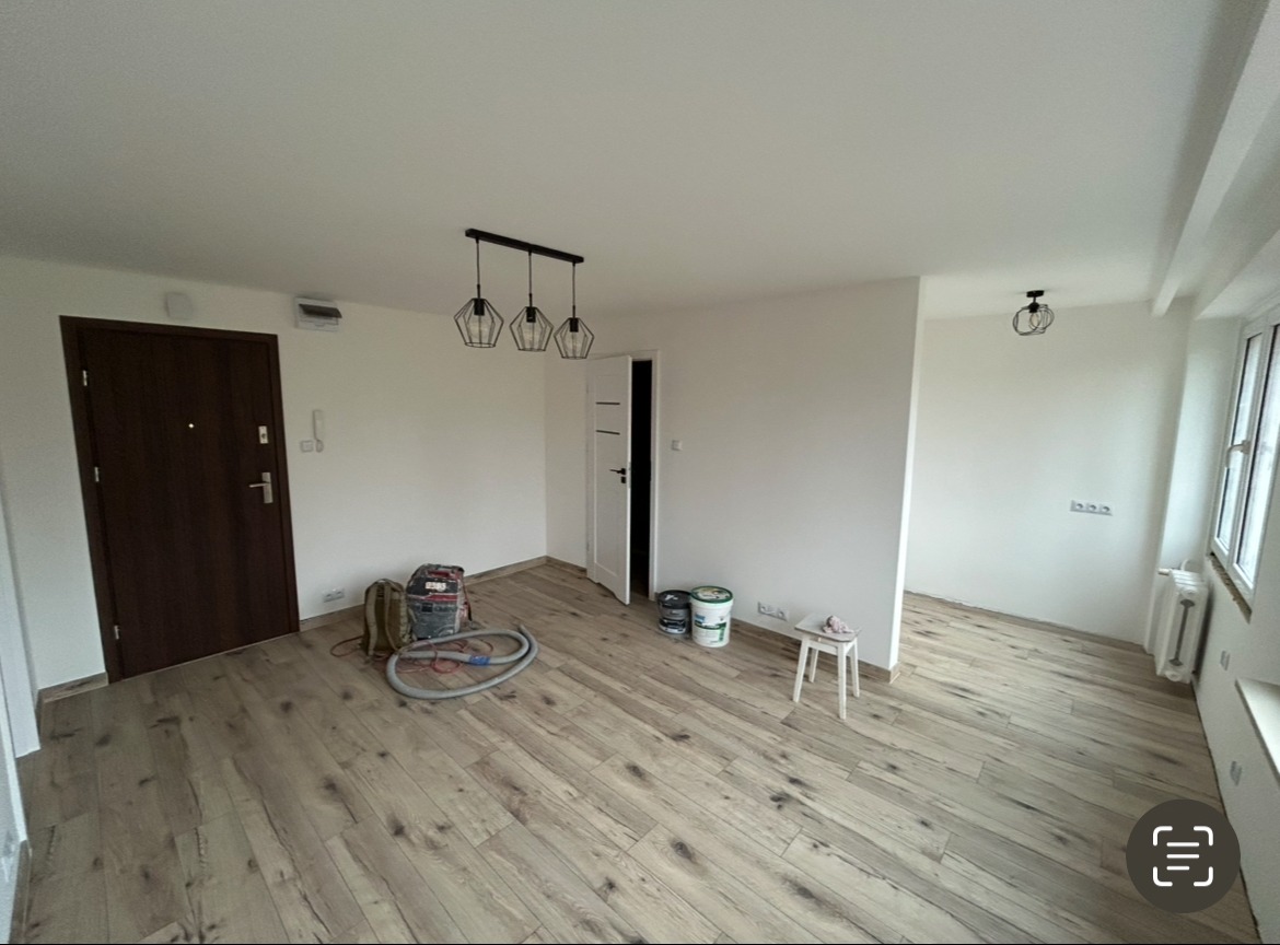 Jasne, odświeżone wnętrze po remoncie: parkiet, pomalowane ściany, nowoczesne lampy. Widoczne akcesoria malarskie i białe drzwi. Minimalistyczny styl.