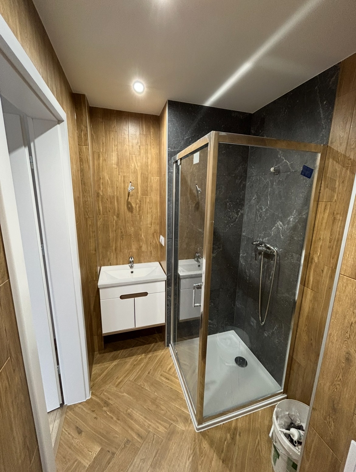 Wykończona łazienka z kabiną prysznicową, umywalką z szafką i drewnopodobnymi płytkami. Nowoczesny design z akcentami drewna i szarości.