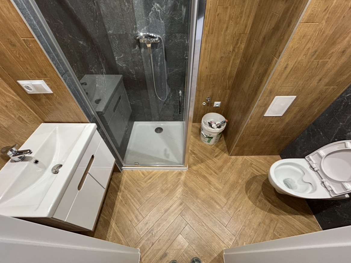 Wykończona łazienka z kabiną prysznicową, umywalką z szafką i toaletą. Uwagę przykuwają płytki imitujące drewno ułożone w jodełkę na podłodze i ścianach.