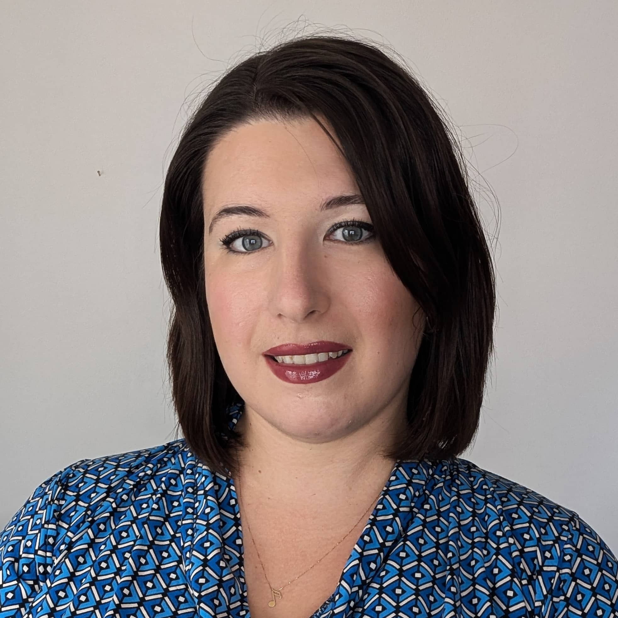 Portret kobiety z krótkimi, ciemnymi włosami, ubranej w niebieską wzorzystą bluzkę i złotą zawieszkę w kształcie nuty. Uśmiech, makijaż z ciemną pomadką.