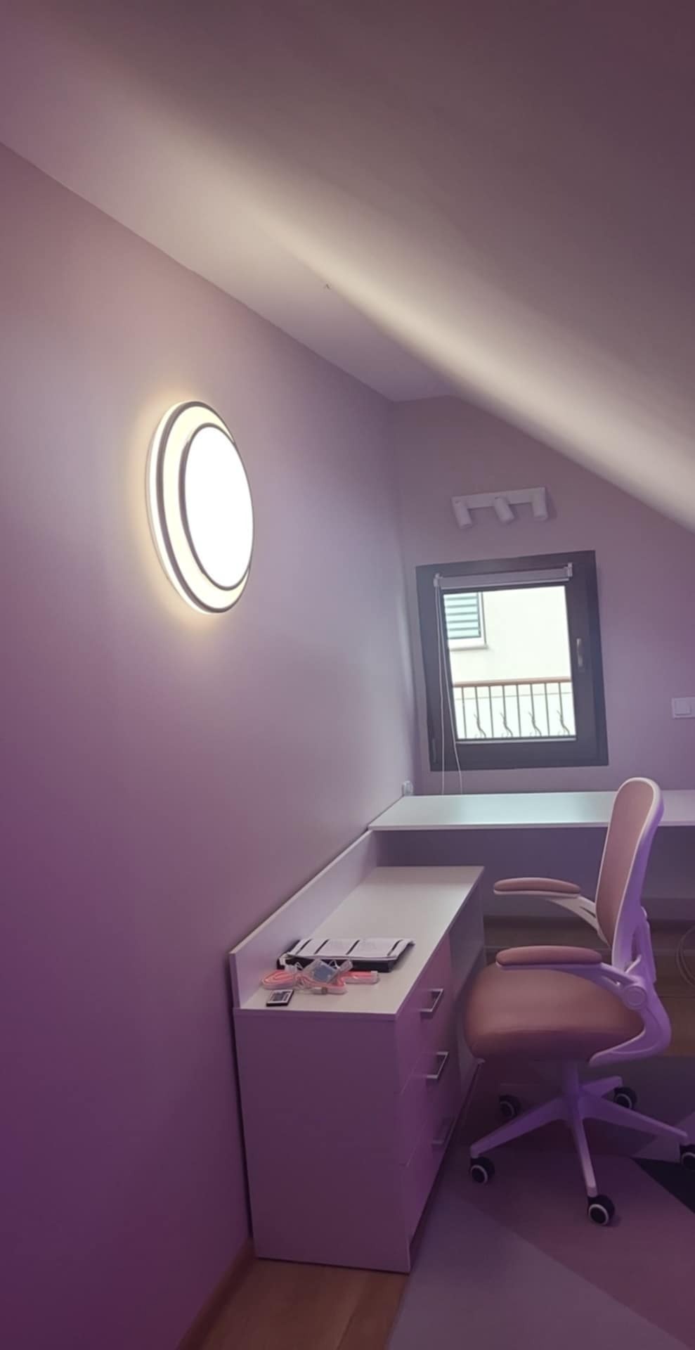 Pokój na poddaszu po odświeżeniu: ściany w odcieniach fioletu, nowoczesne oświetlenie ścienne i biurko z krzesłem. Widok na okno dachowe i skos.