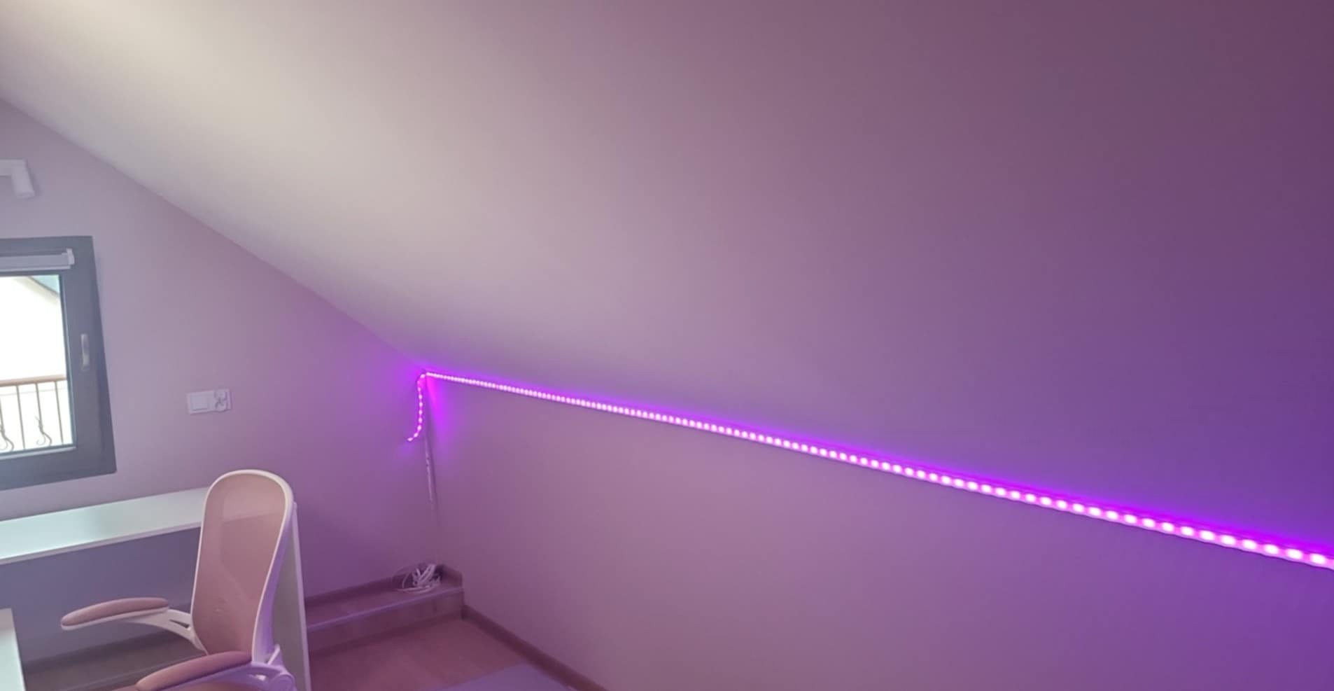 Pokój na poddaszu z różową ścianą i oświetleniem LED, biurko z oknem i krzesłem. Nietypowe oświetlenie nadaje pomieszczeniu nowoczesny charakter.