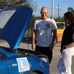 Auto Szkoła Premium - Instruktor z kursantką przy niebieskim aucie z otwartą maską i naklejką 'L' w Łodzi. Żółty samochód szkoły jazdy w tle.