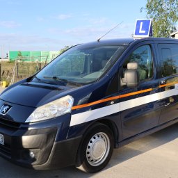Auto Szkoła Premium - Niebieski Peugeot z pomarańczowymi pasami i literą 'L' na dachu, zaparkowany na placu manewrowym szkoły jazdy, w tle widoczna infrastruktura drogowa.